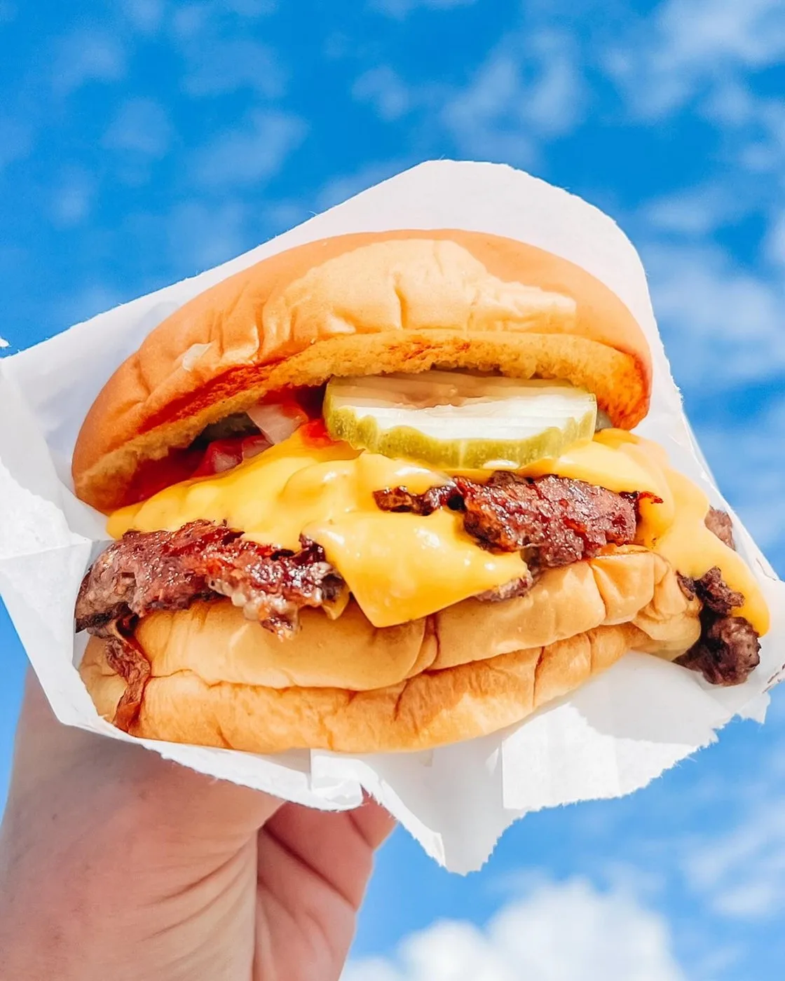 A double smash burger with melted American cheese and a dill chip, held up against a blue sky.