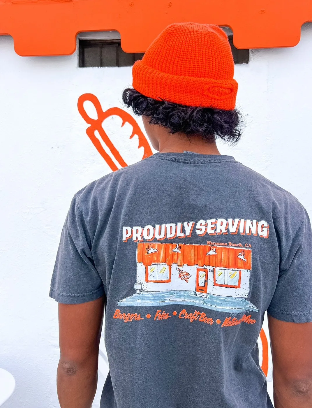 Someone in an orange beanie wearing a Proudly Serving t-shirt printed with the Hermosa Beach storefront.