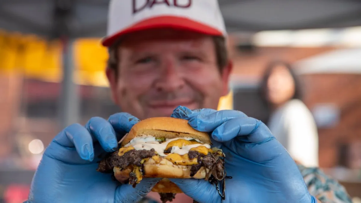 A man in a "DAD" cap holding up a double cheeseburger in gloved hands.