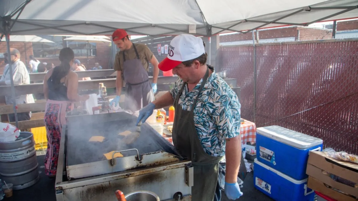 A cook pressing patties on a flattop under a pop-up canopy, steam coming off the griddle.