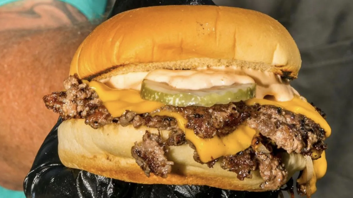 A gloved hand holding a double smash cheeseburger dressed with sauce and a pickle slice.