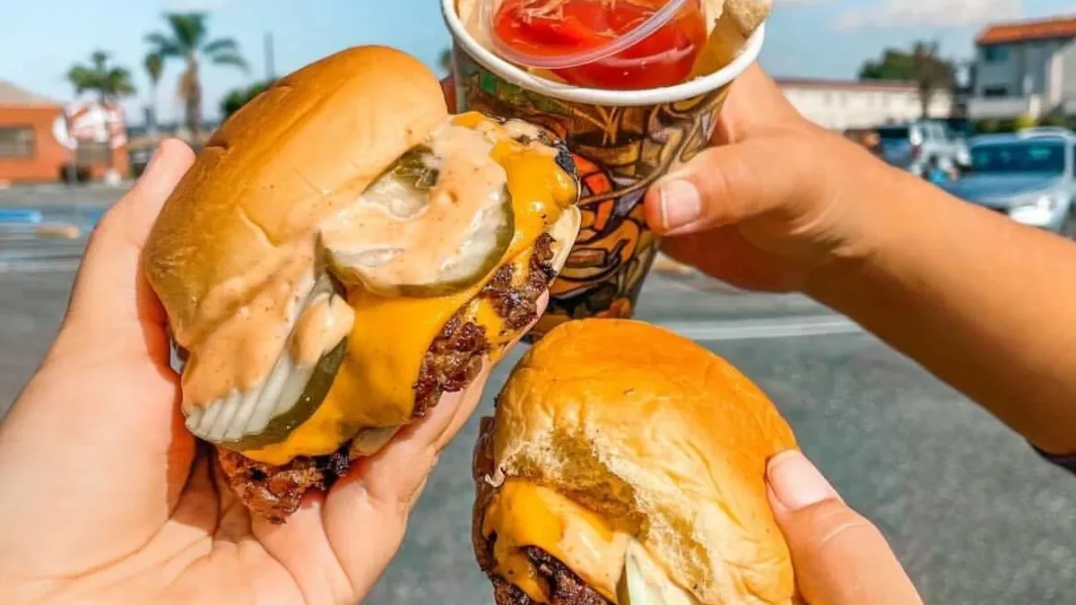 Two hands holding smash cheeseburgers and a third holding a soda, out in a sunlit lot.